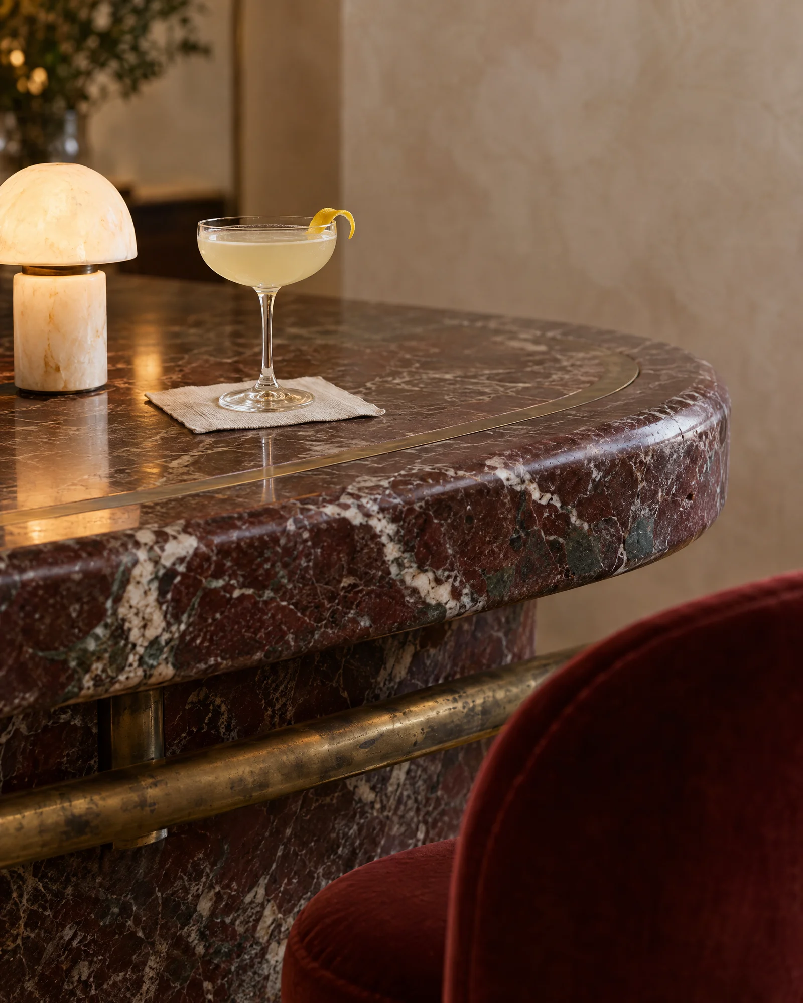 Concept hotel lobby bar detail: a honed Rosso Levanto marble bar edge with antique bronze inlay and rail, a cocktail coupe, an alabaster lamp and a red velvet stool